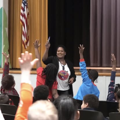 Creator of Mya, E. Moore leading an interactive school assembly with excited children raising hands